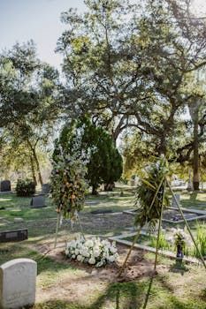 A grave in a cemetery decorated with floral arrangements and standing wreaths under large trees on a sunny day.
