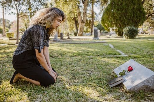 A person with curly hair kneels on the grass in a cemetery before a headstone marked with a single red rose and a book.