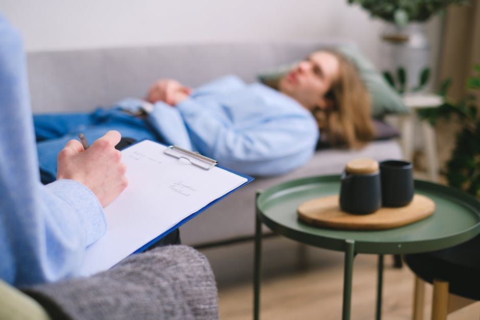 A person taking notes on a clipboard while a patient lies on a couch during a therapy session.