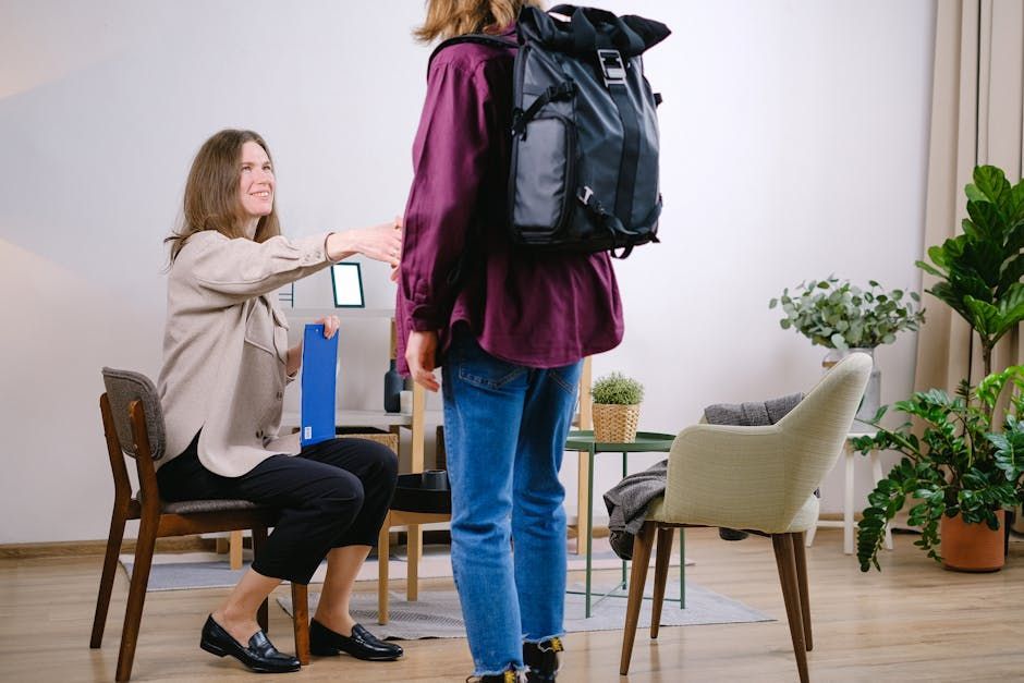 A person seated holds a blue folder and reaches out to shake hands with a standing person wearing a large black backpack.
