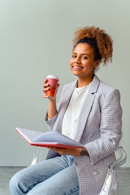 A person sitting in a transparent chair, holding a coffee cup and an open notebook, smiling against a neutral background.