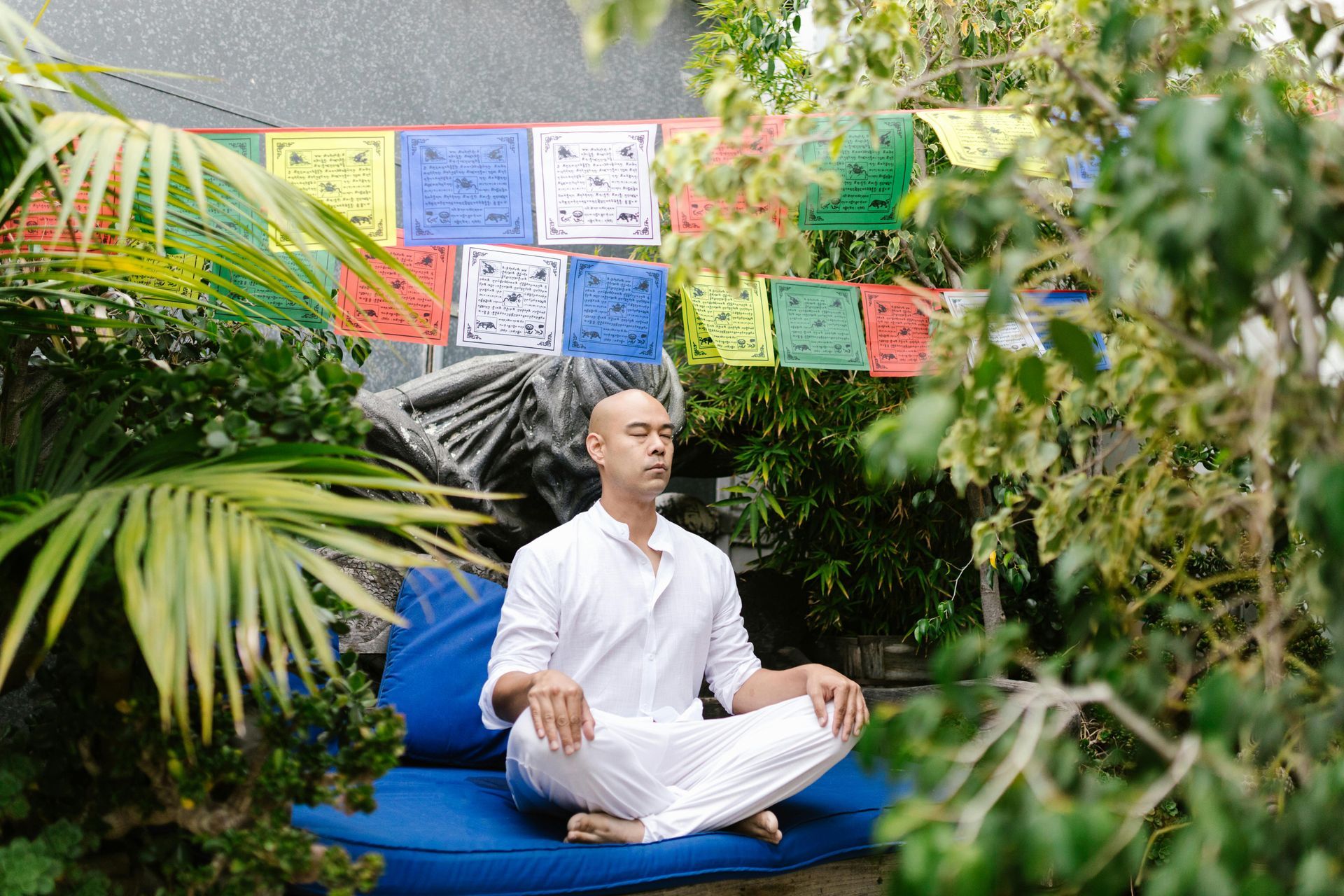 Person in white clothes meditating cross-legged on a blue cushion in a lush garden with colorful prayer flags.