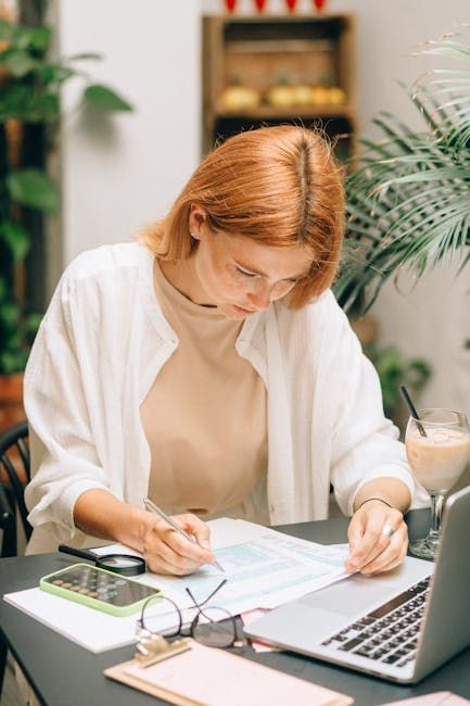 A person works at a desk, writing on documents near a laptop, glasses, and a drink, with plants in the background.