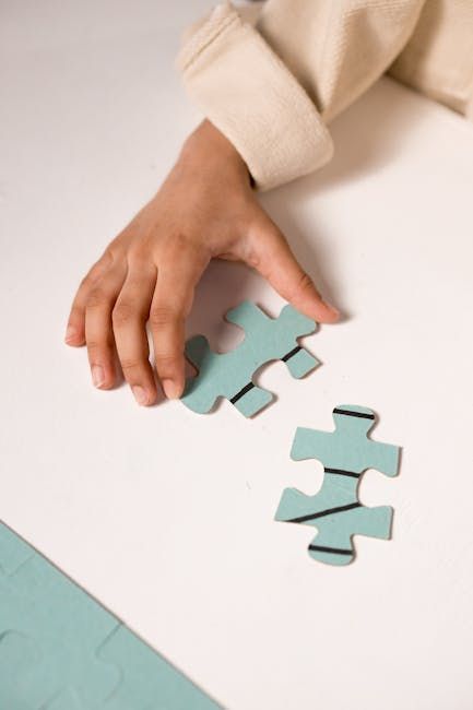 A person's hand reaches for a light blue puzzle piece on a white surface near another matching piece with black lines.