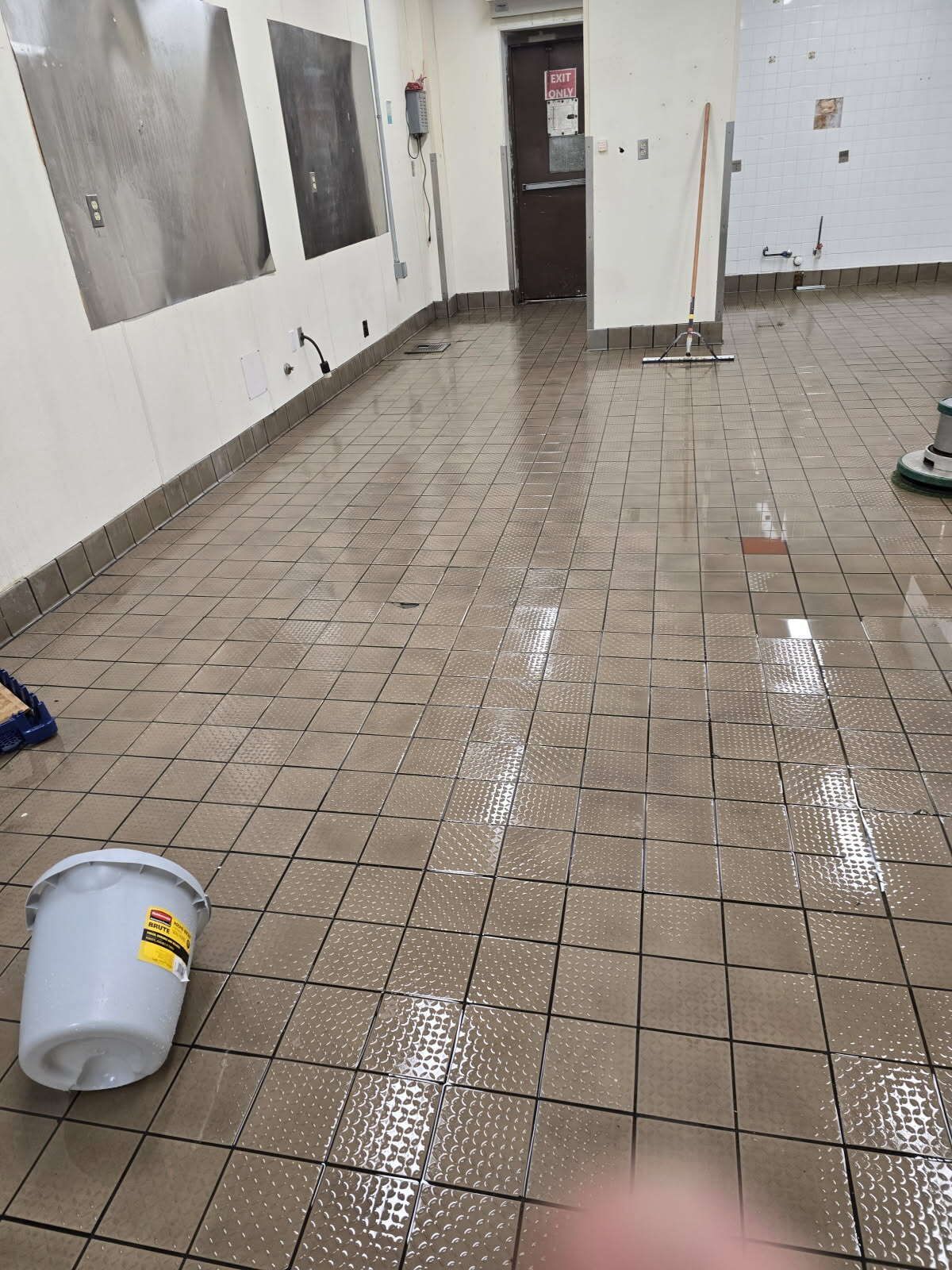Flooded, tiled floor in a commercial kitchen with a bucket, cleaning supplies, and a doorway in the background.