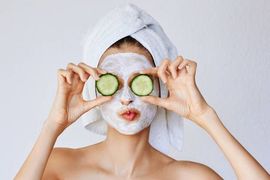 A person wearing a white face mask and hair towel holds cucumber slices over their eyes while making a playful expression.