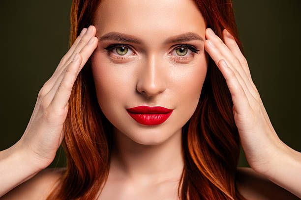 Close-up of a red-haired woman with bold red lipstick, touching her temples against a dark background