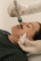 A professional wearing pink gloves uses a small syringe to administer a cosmetic injection near a smiling person's eye.