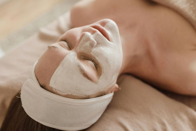 A person lying down with a facial mask applied, wearing a white headband, resting on a beige surface.