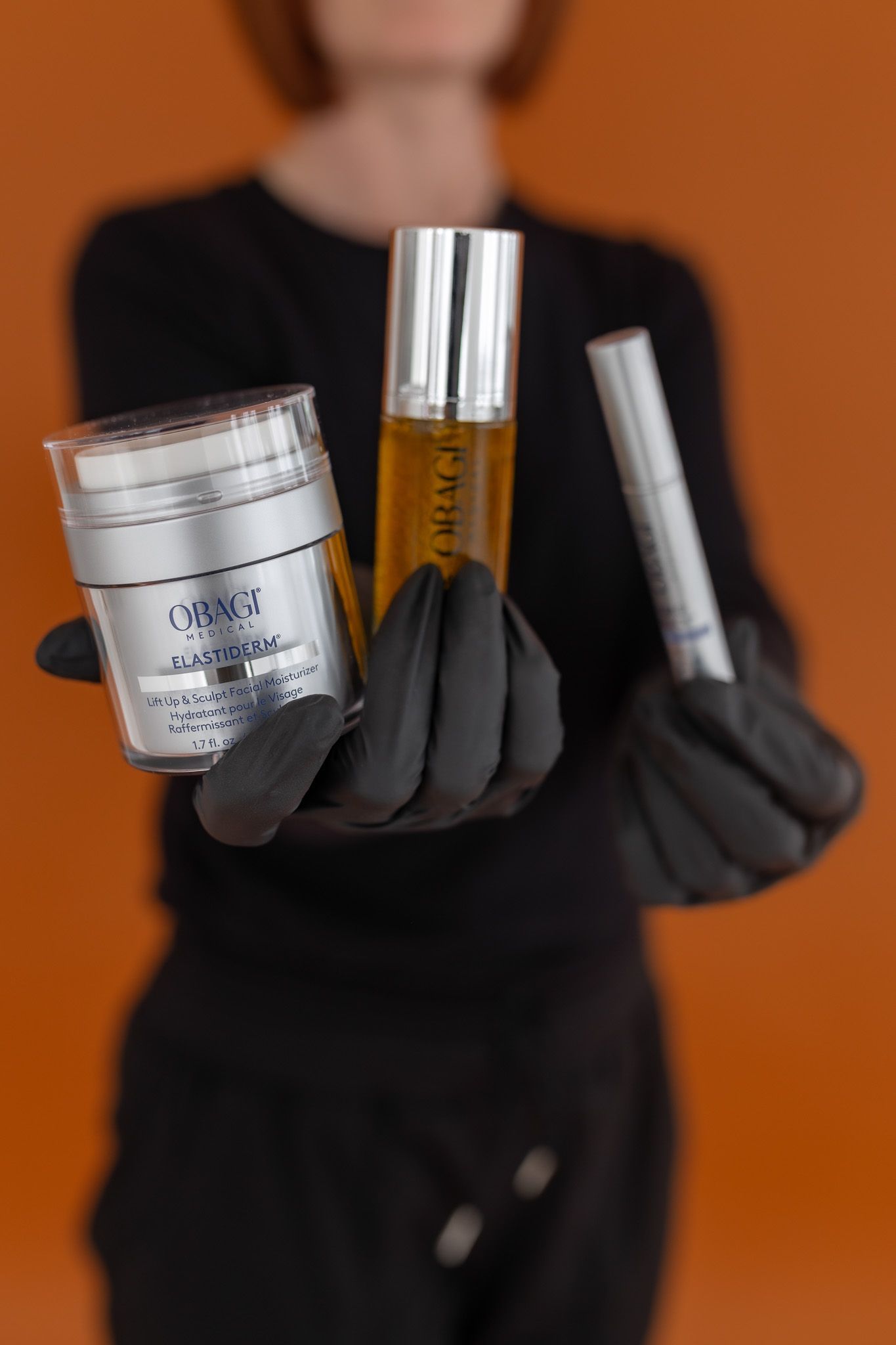 Gloved person holding three cosmetic products against an orange background