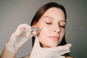 A person receives a cosmetic facial injection from a professional wearing white gloves against a gray background.
