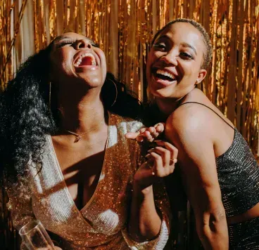 Two people laugh while posing in sequined outfits against a metallic gold fringe backdrop, one holding a glass of wine.