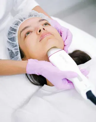 Person with a white face mask and towel wrap holding cucumber slices over their eyes.