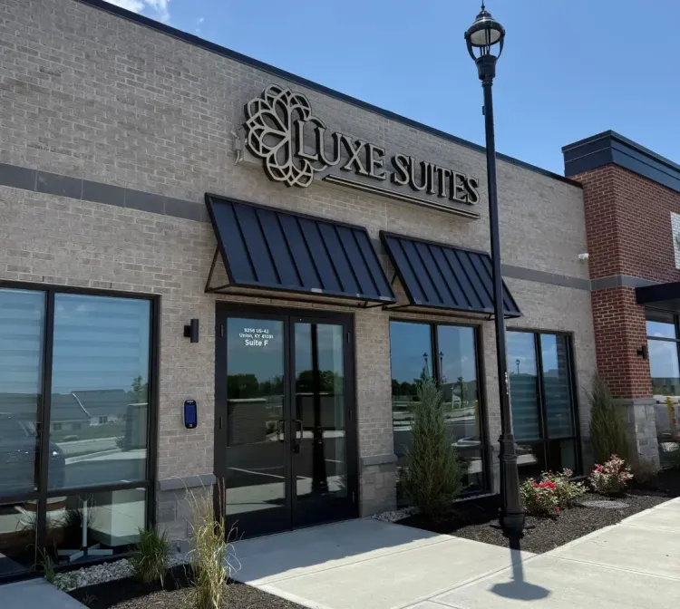 The storefront of Luxe Suites with beige brick walls, black metal awnings, glass double doors, and a street lamp.