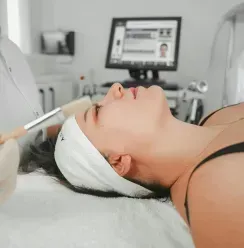 Person receiving a facial treatment with a handheld device in a spa setting, lying on a treatment bed.