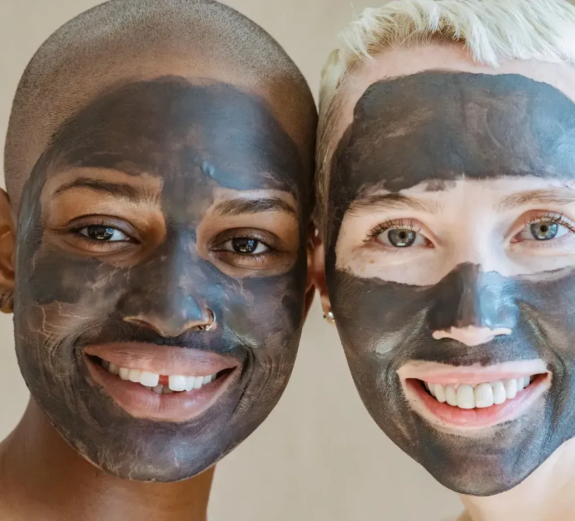Two people with short hair wearing charcoal face masks, smiling at the camera against a neutral background.
