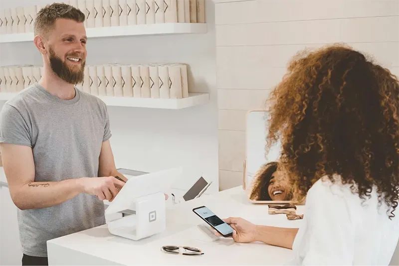 A customer paying with a smartphone at a retail checkout counter while a store associate operates the point-of-sale system.