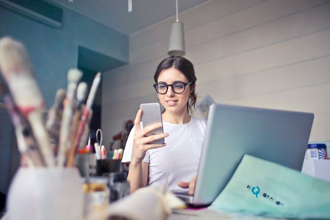 Woman in glasses using phone and laptop, brushes in background.