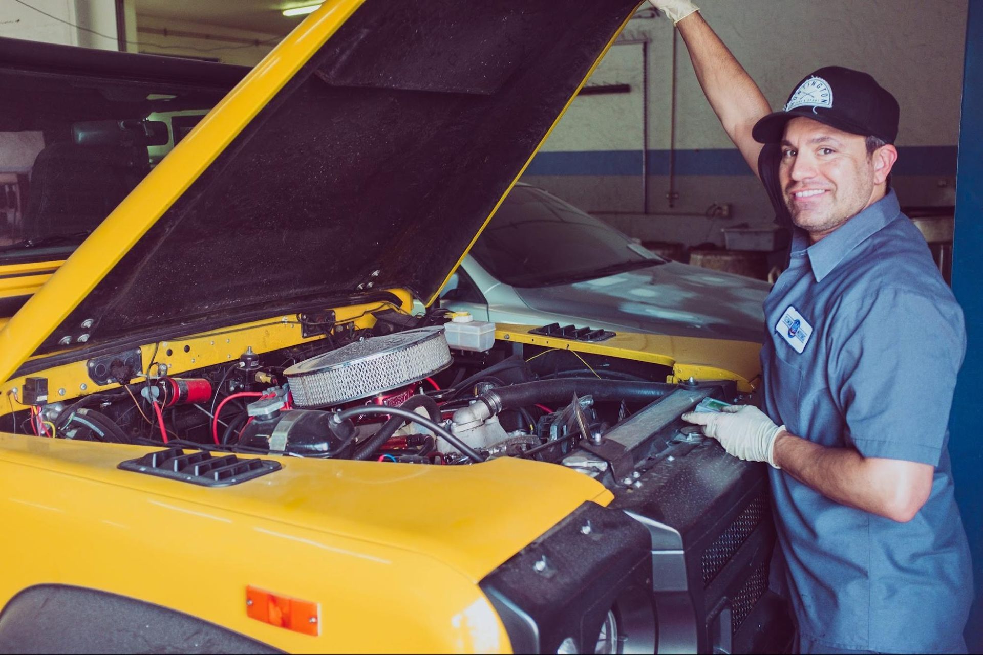 Mechanic smiling beside a yellow car with its hood open in a garage