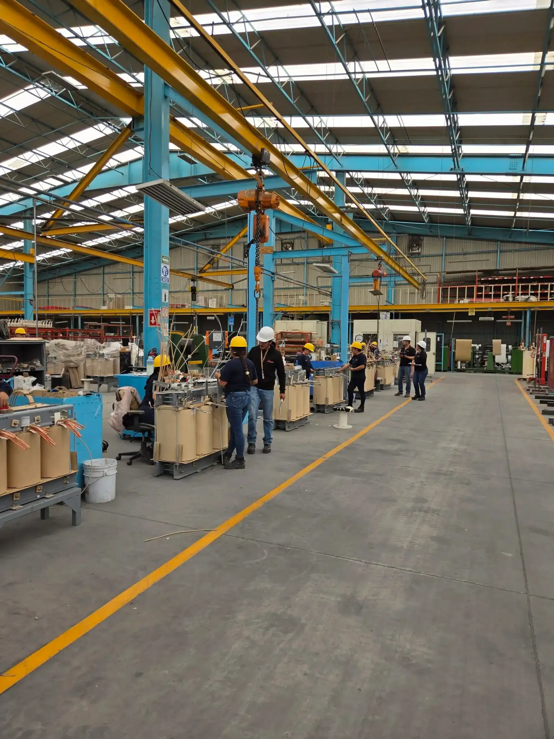 People working on an assembly line in a large warehouse with blue support columns and overhead cranes.