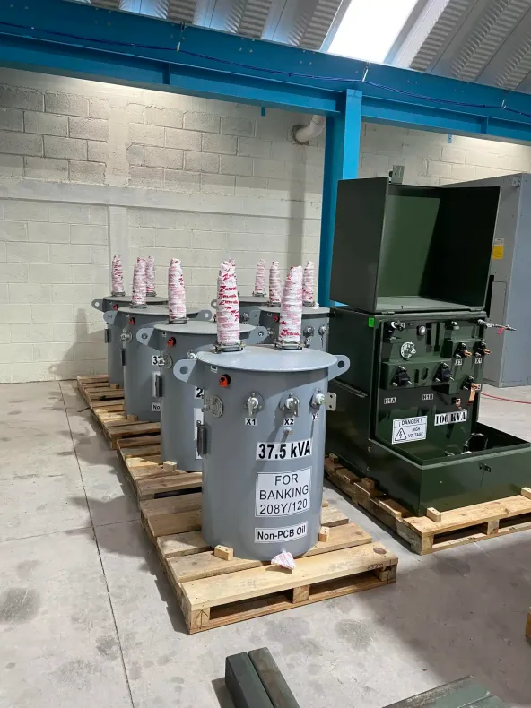 Electrical transformers on wooden pallets in an industrial warehouse, with one open green unit and gray units nearby