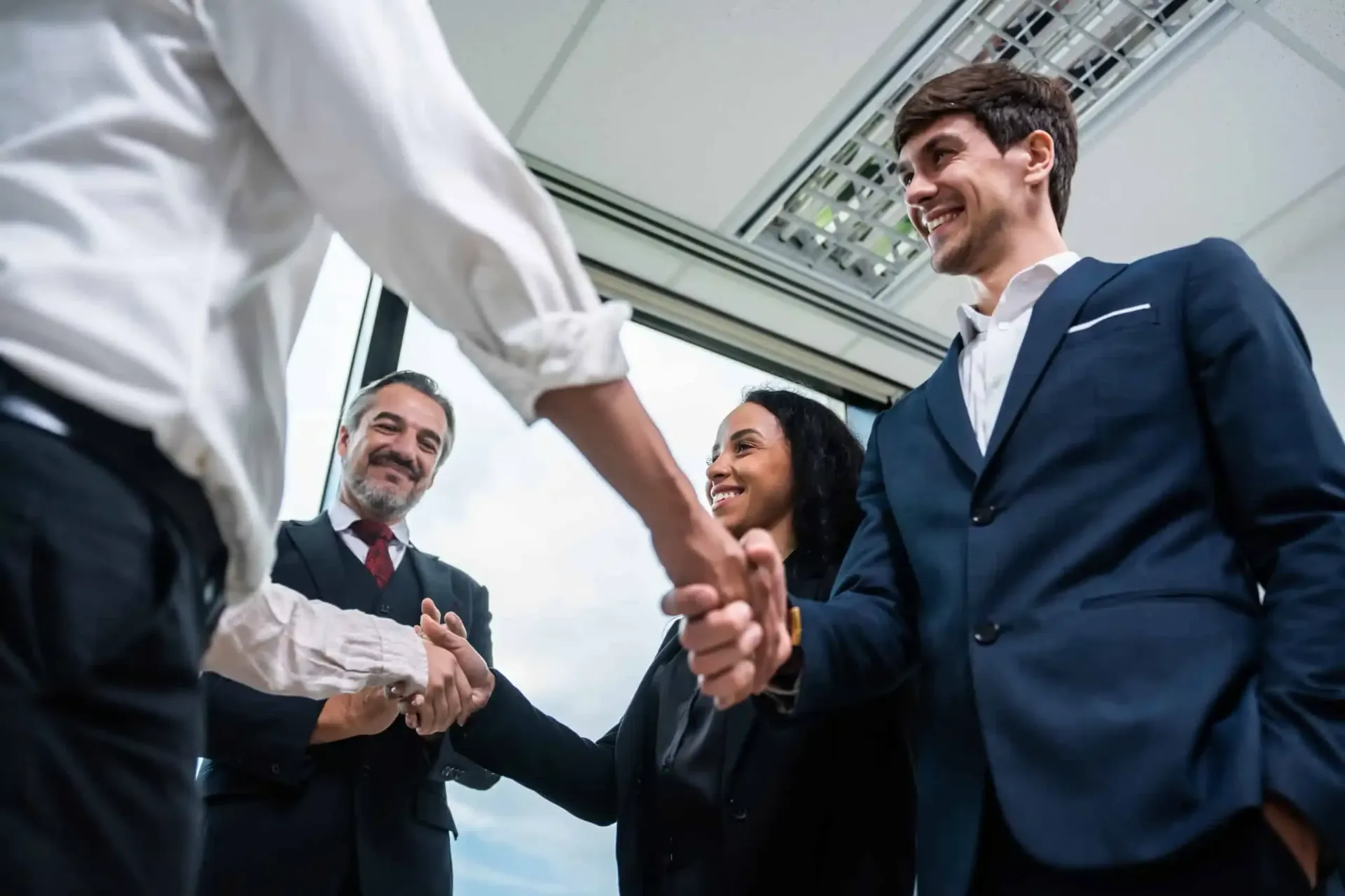 Businesspeople shaking hands in a bright office, with smiling colleagues in suits nearby.