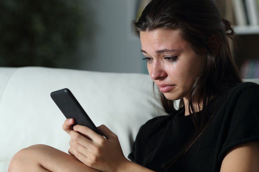 A woman is crying while sitting on a couch looking at her cell phone.