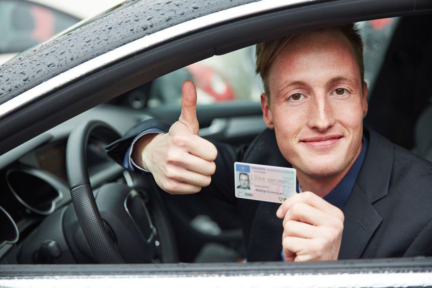A man is sitting in a car holding a driving license and giving a thumbs up.