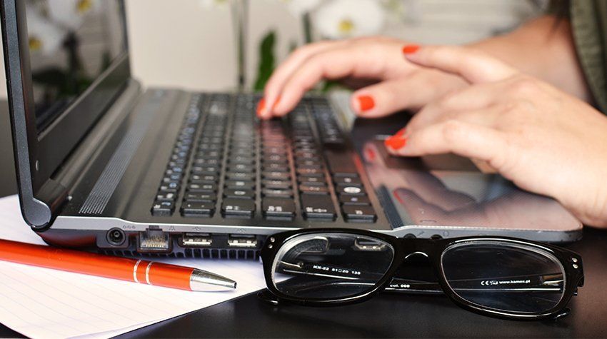 A woman is typing on a laptop next to a pair of glasses and a pen