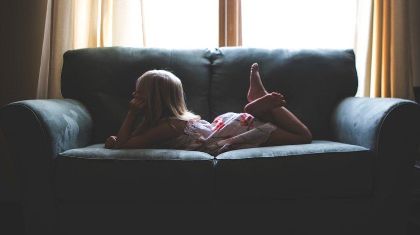 A little girl is laying on a couch in front of a window.