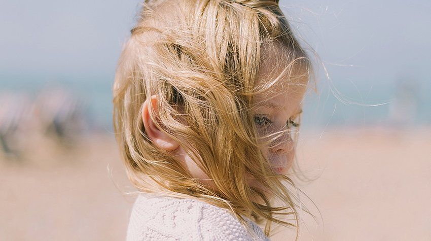 A little girl is standing on a beach with her hair blowing in the wind.