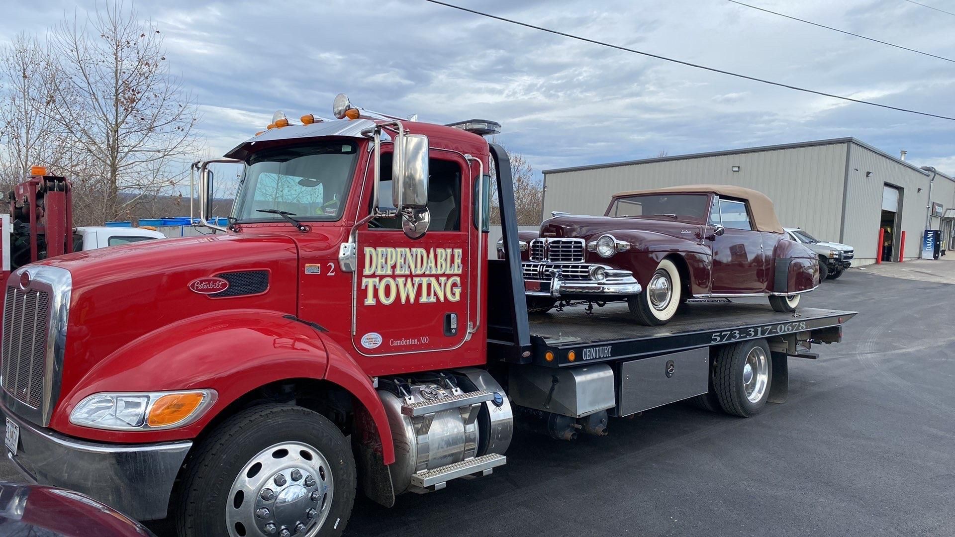 Red towing truck carrying a maroon classic convertible on a flatbed in a parking lot | Dependable Automotive