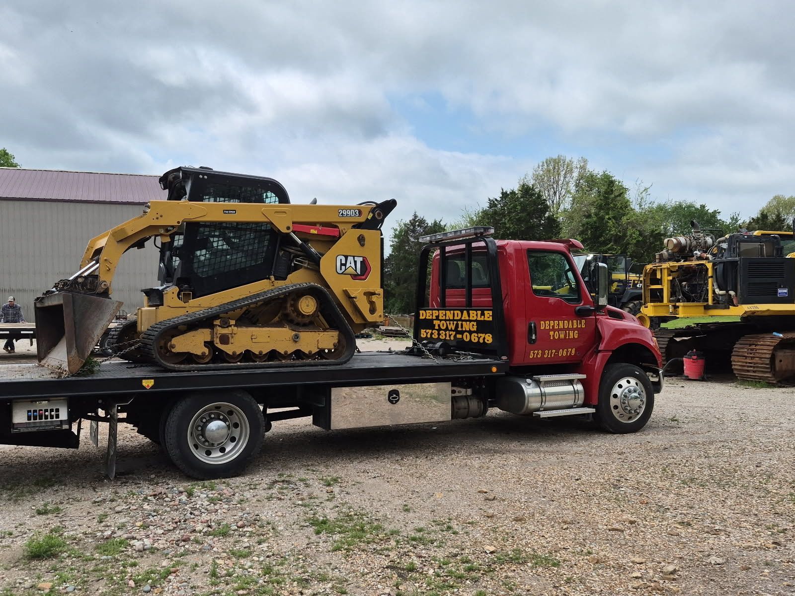 Red tow truck carrying a yellow CAT skid steer on a flatbed in a gravel lot | Dependable Automotive