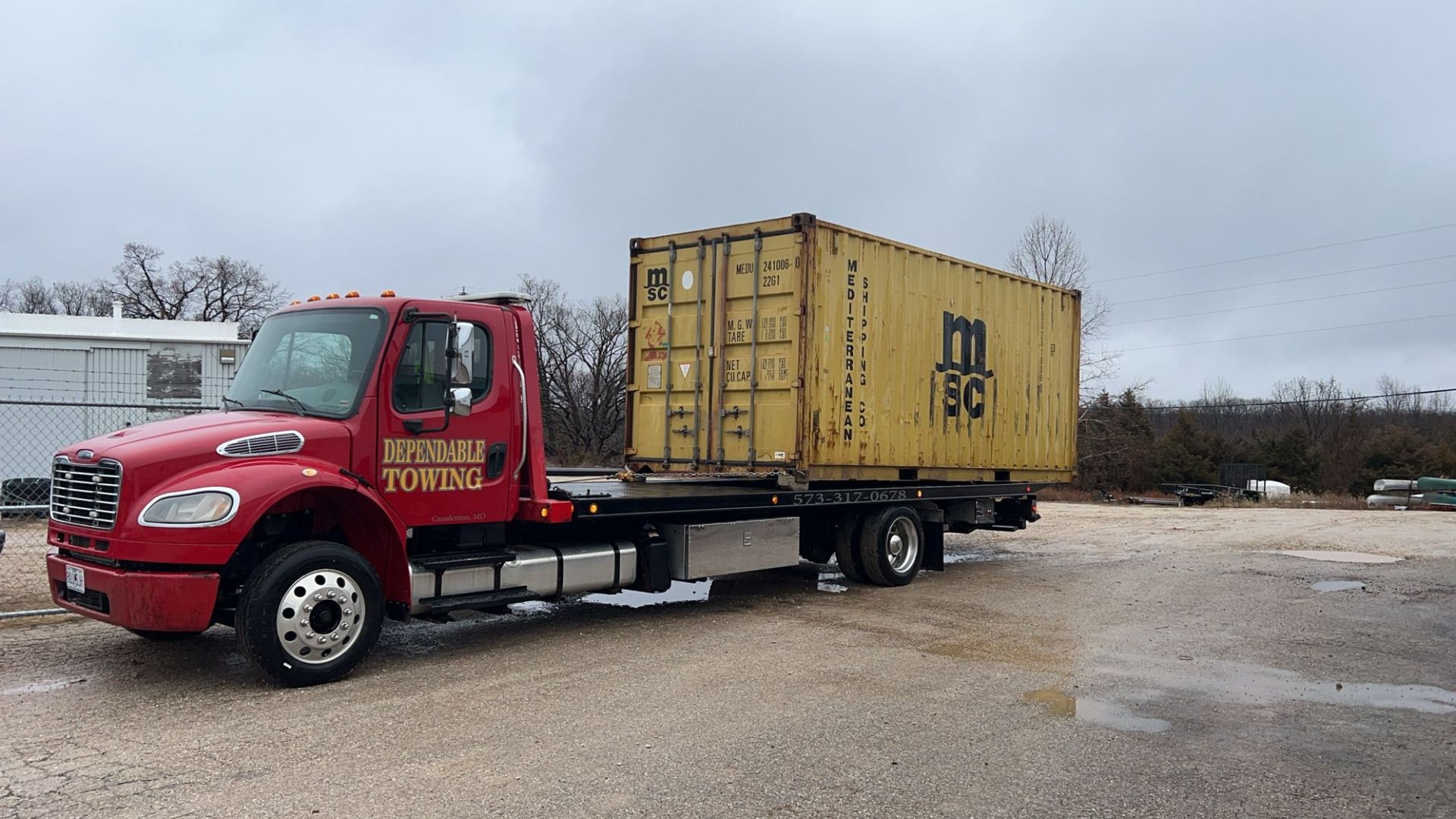 Red flatbed truck hauling a yellow shipping container on a gravel lot under cloudy skies | Dependable Automotive