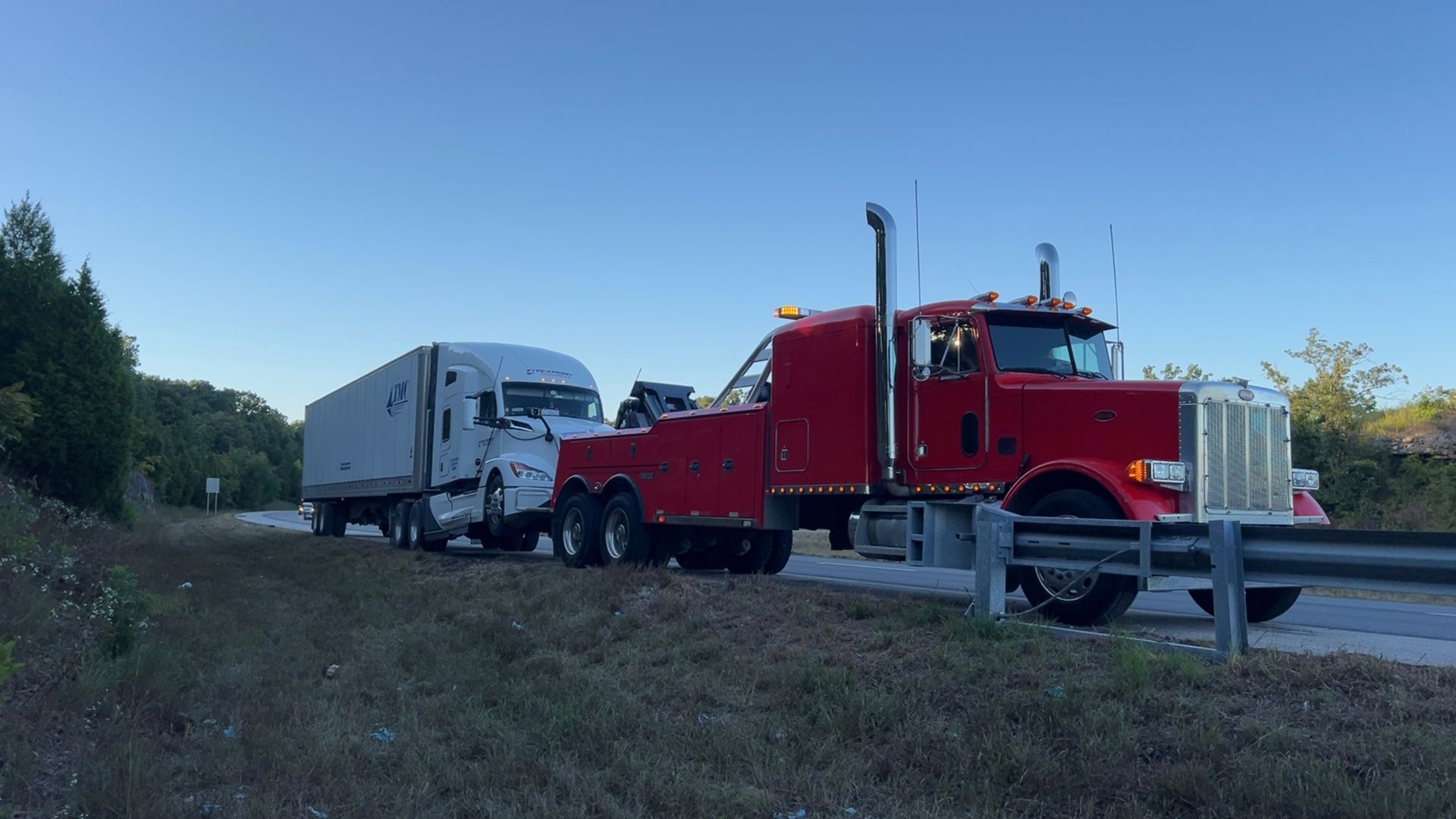 Red tow truck pulling a white semi-trailer on a roadside at dusk | Dependable Automotive