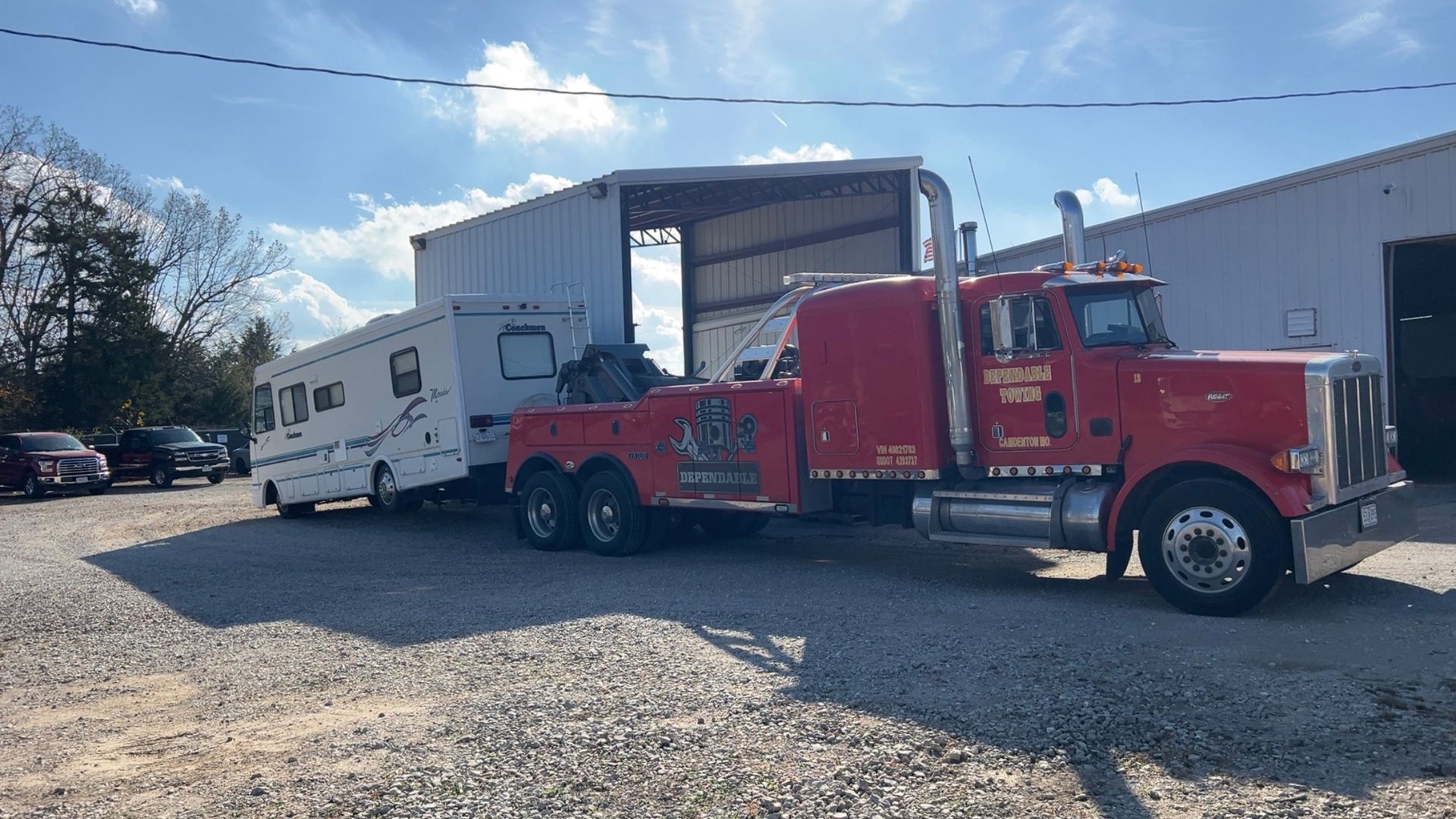 Red tow truck hauling a white RV beside an open garage on a gravel lot. | Dependable Automotive