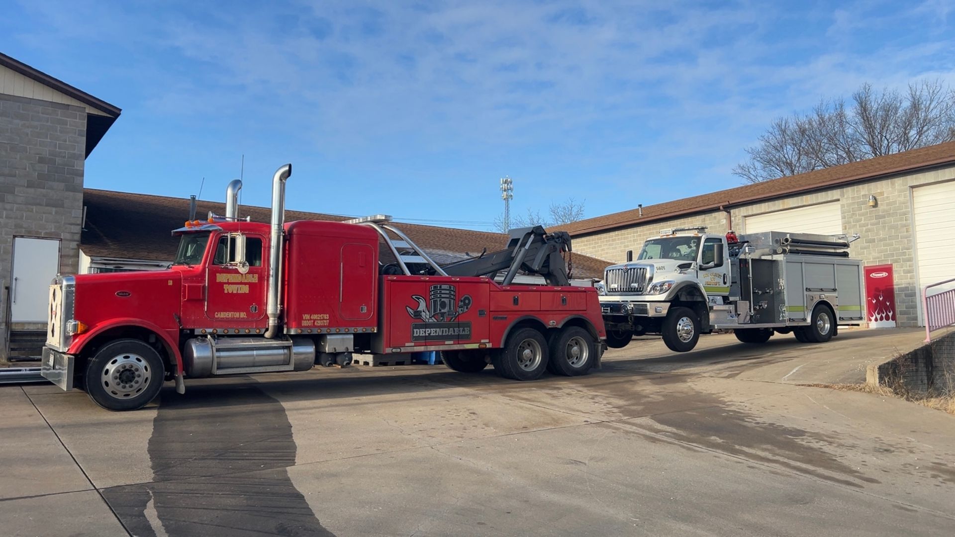Red tow truck hauling a white pickup and trailer in a parking lot under a clear blue sky | Dependable Automotive