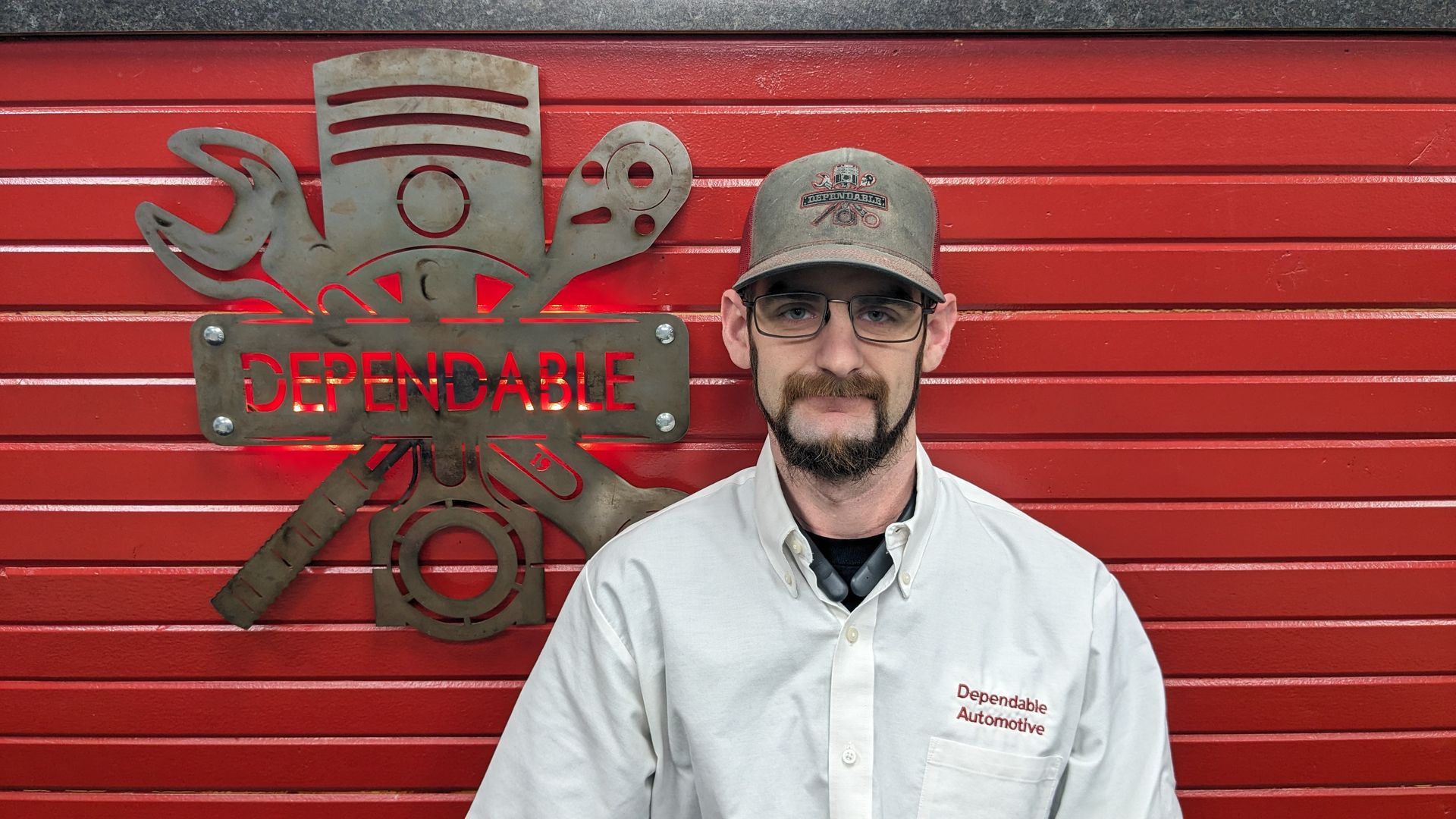 Man standing in front of a red metal wall with a sign and crossed tools logo | Dependable Automotive