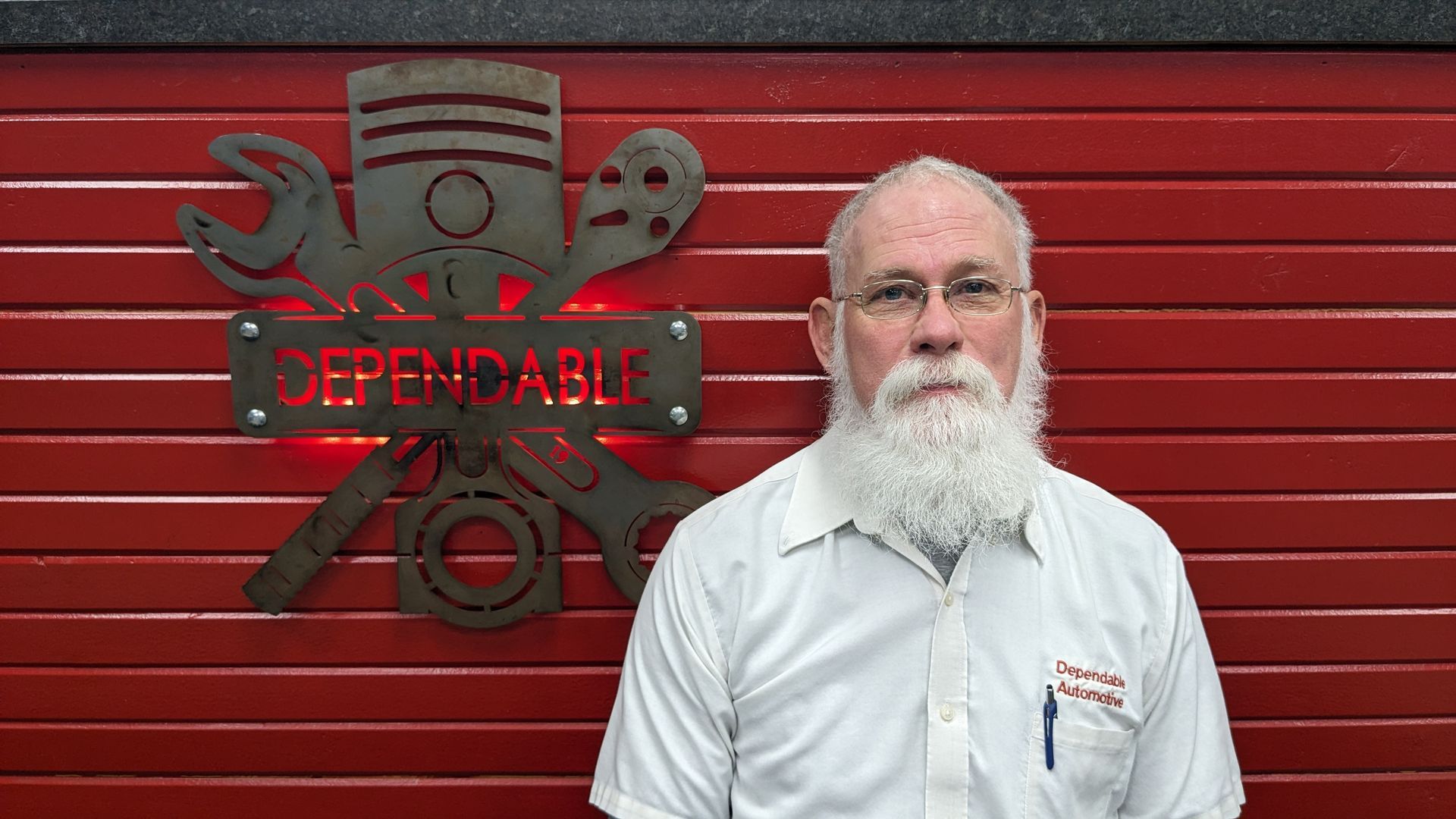 Man in white shirt standing before a red wall with a black CrossFit-style logo | Dependable Automotive
