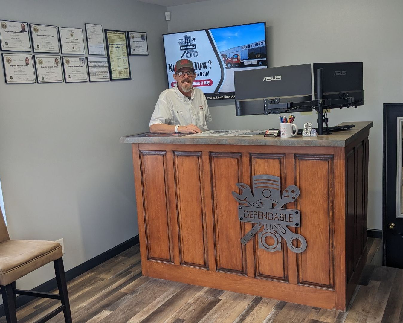 Person seated behind a wooden front desk in a bright office reception area, with monitors and framed certificates nearby | Dependable Automotive