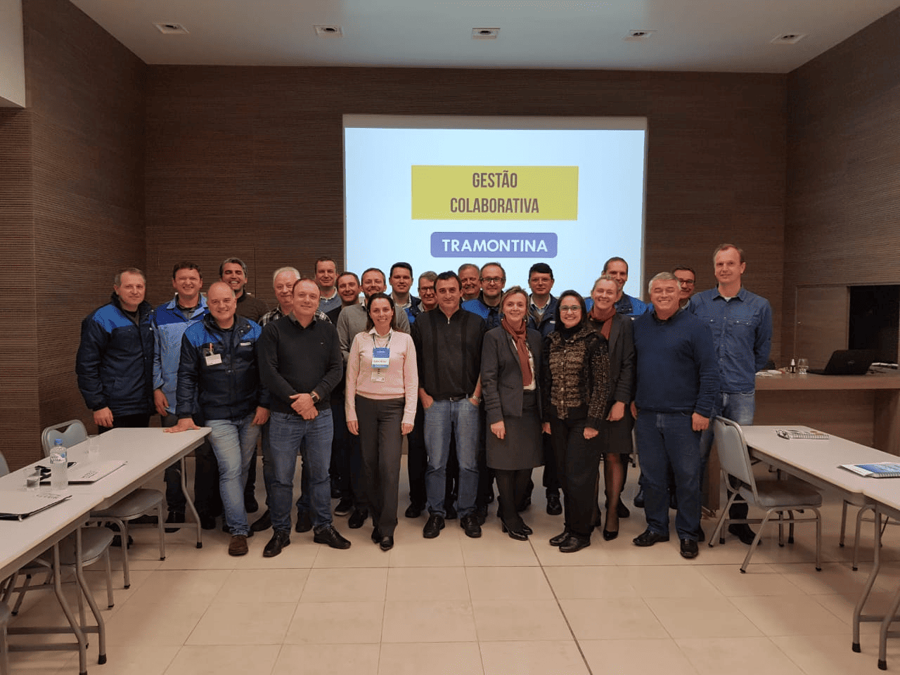 Grupo de pessoas posando em uma sala de conferências em frente a uma tela de projeção com slides de apresentação.