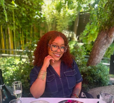Smiling person seated outdoors at a table with a drink, surrounded by lush greenery.