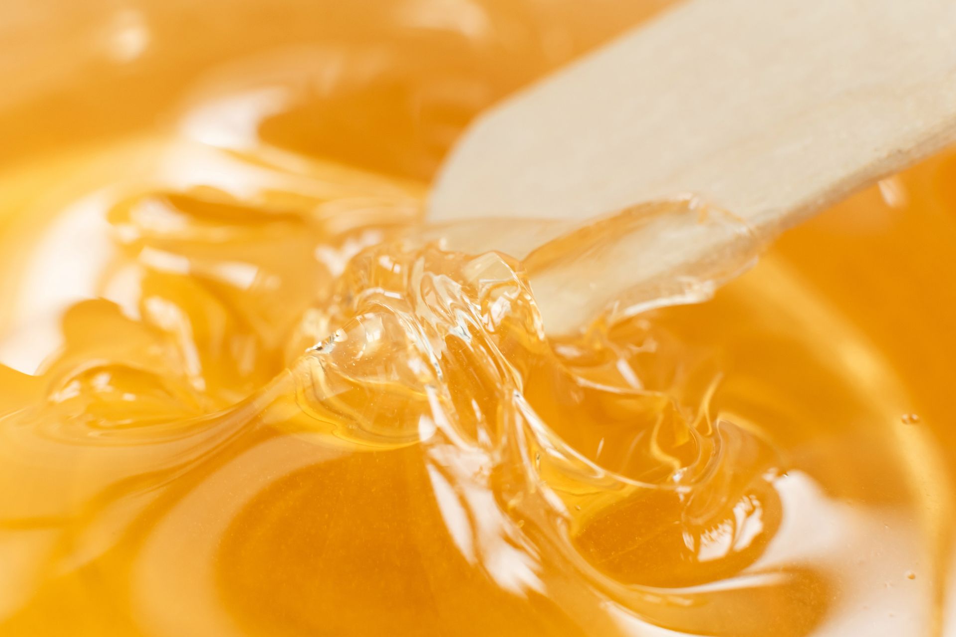 A wooden applicator tool stirring a thick, amber-colored sugar wax or honey mixture.