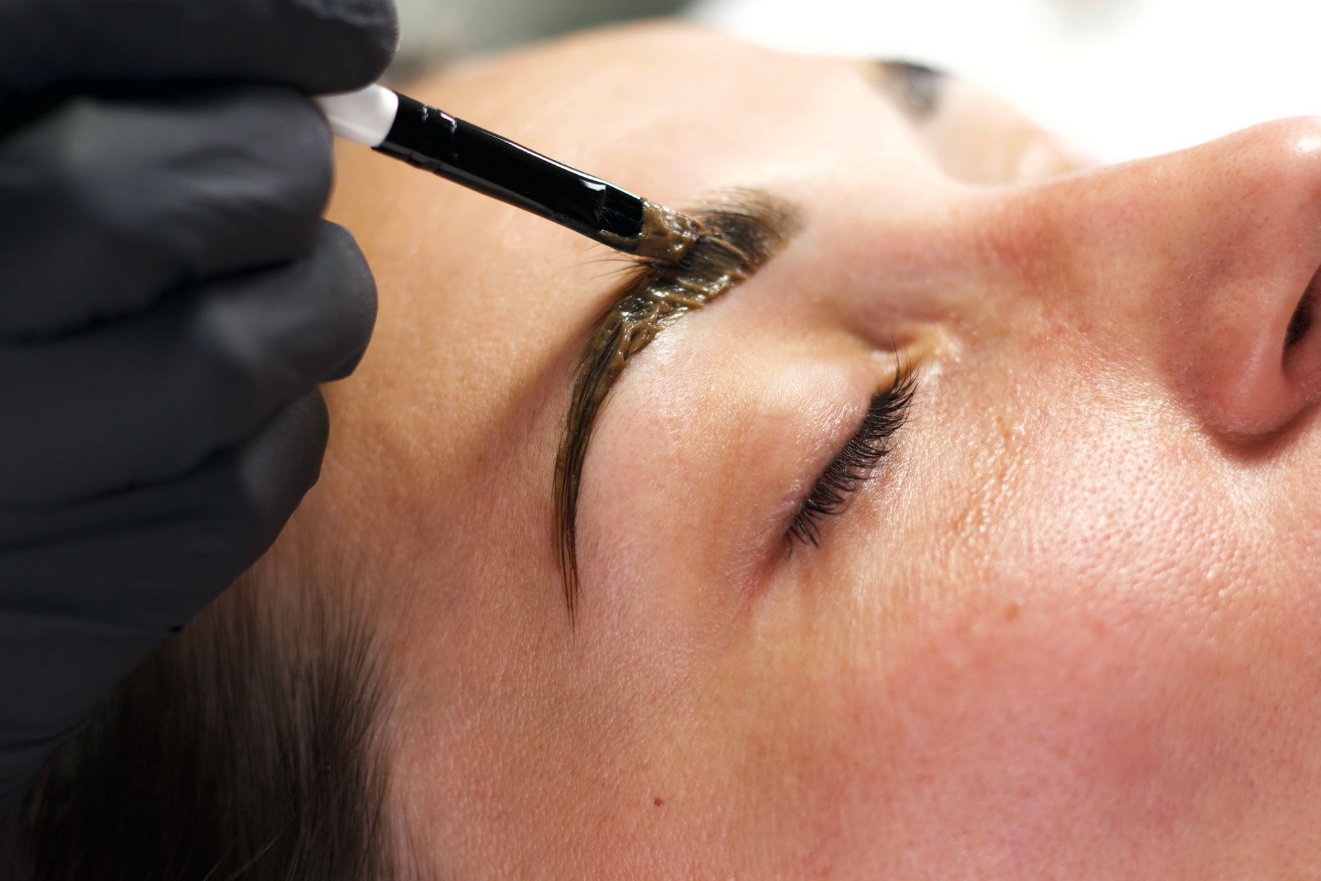 Gloved hand brushing dark dye onto a woman's eyebrow during a beauty treatment