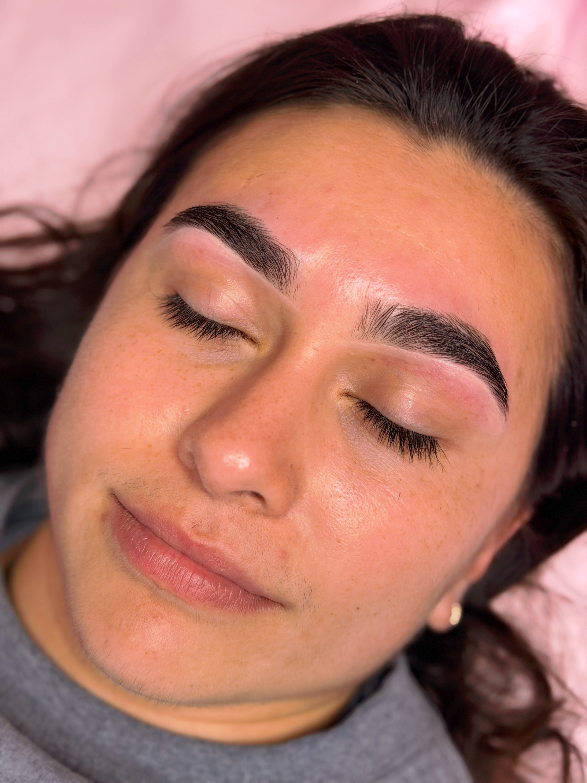 Close-up of a person lying down with eyes closed and neatly shaped brows on a pink background