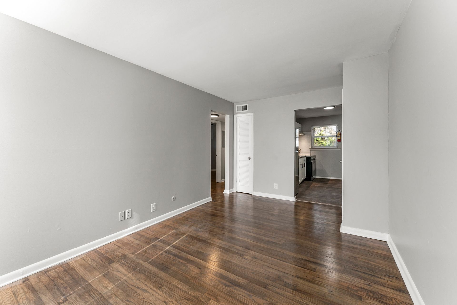 Empty room with gray walls, hardwood floors, and an open doorway to a kitchen area.