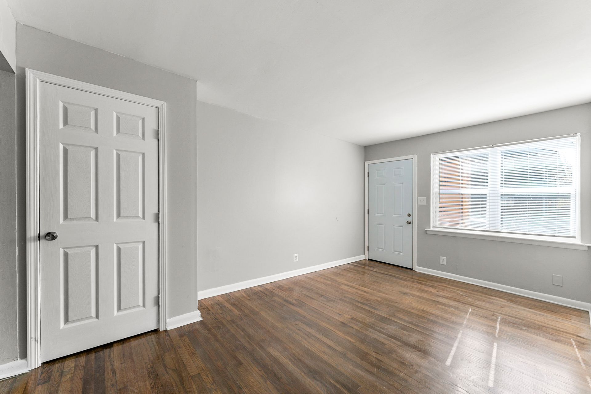 Empty gray living room with hardwood floors, white doors, and a window with blinds