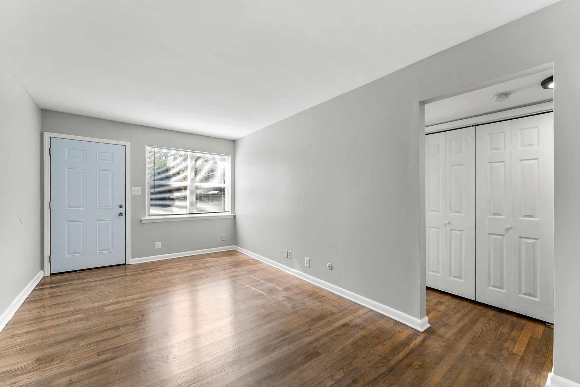 Empty room with hardwood floors, gray walls, blue closet door, and a window with blinds.