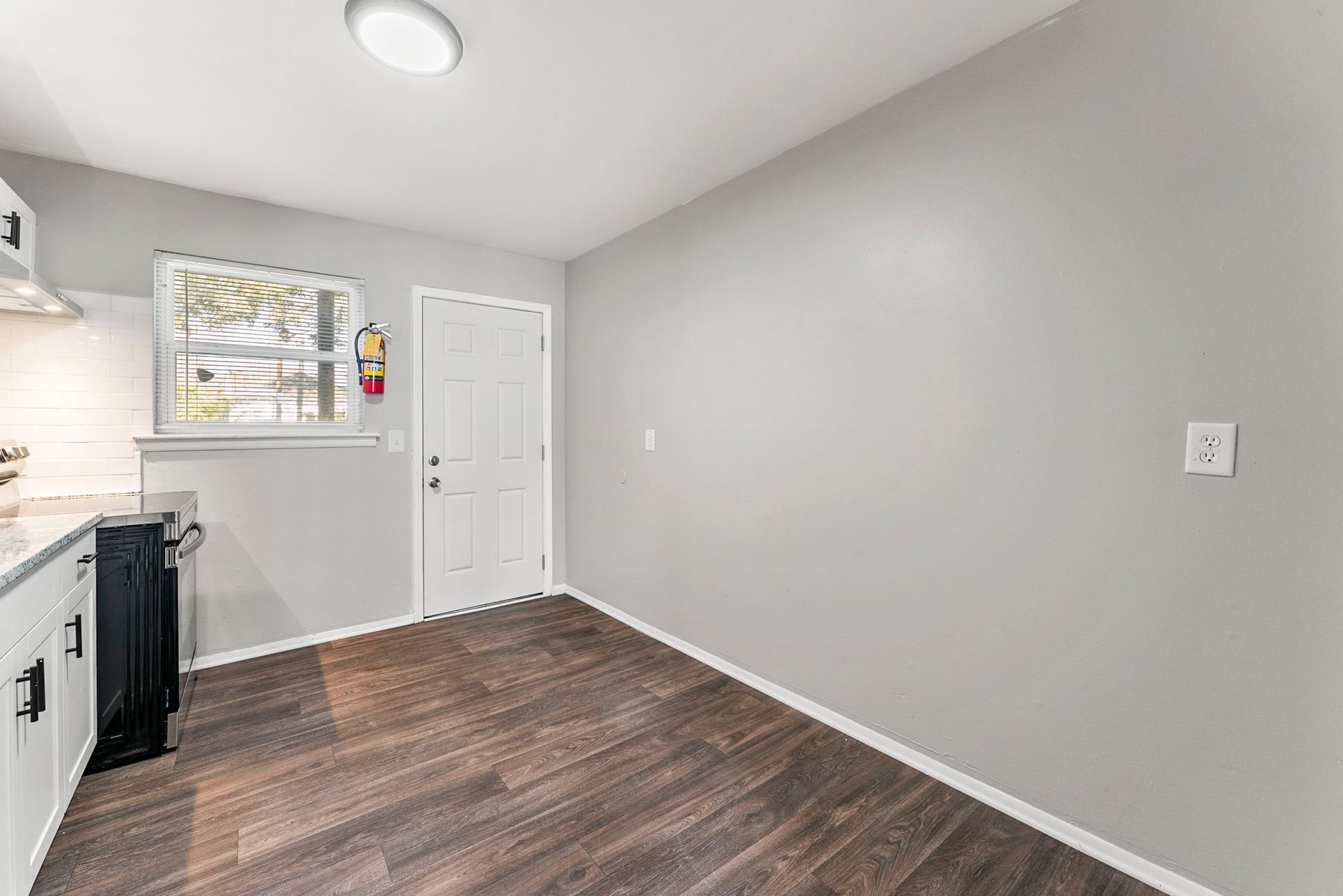 Empty kitchen with gray walls, white cabinets, and wood-look flooring, viewed toward a door and window.