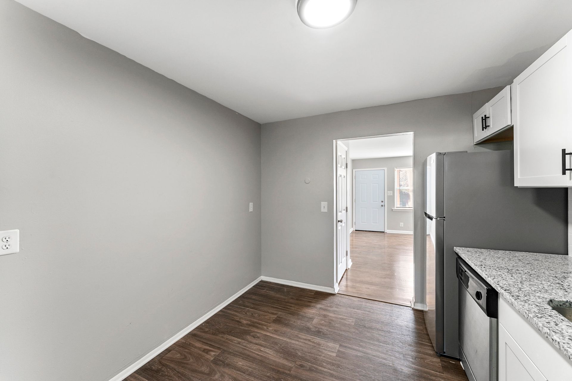 Empty kitchen with gray walls, dark wood floor, white cabinets, and a doorway to a bright room.